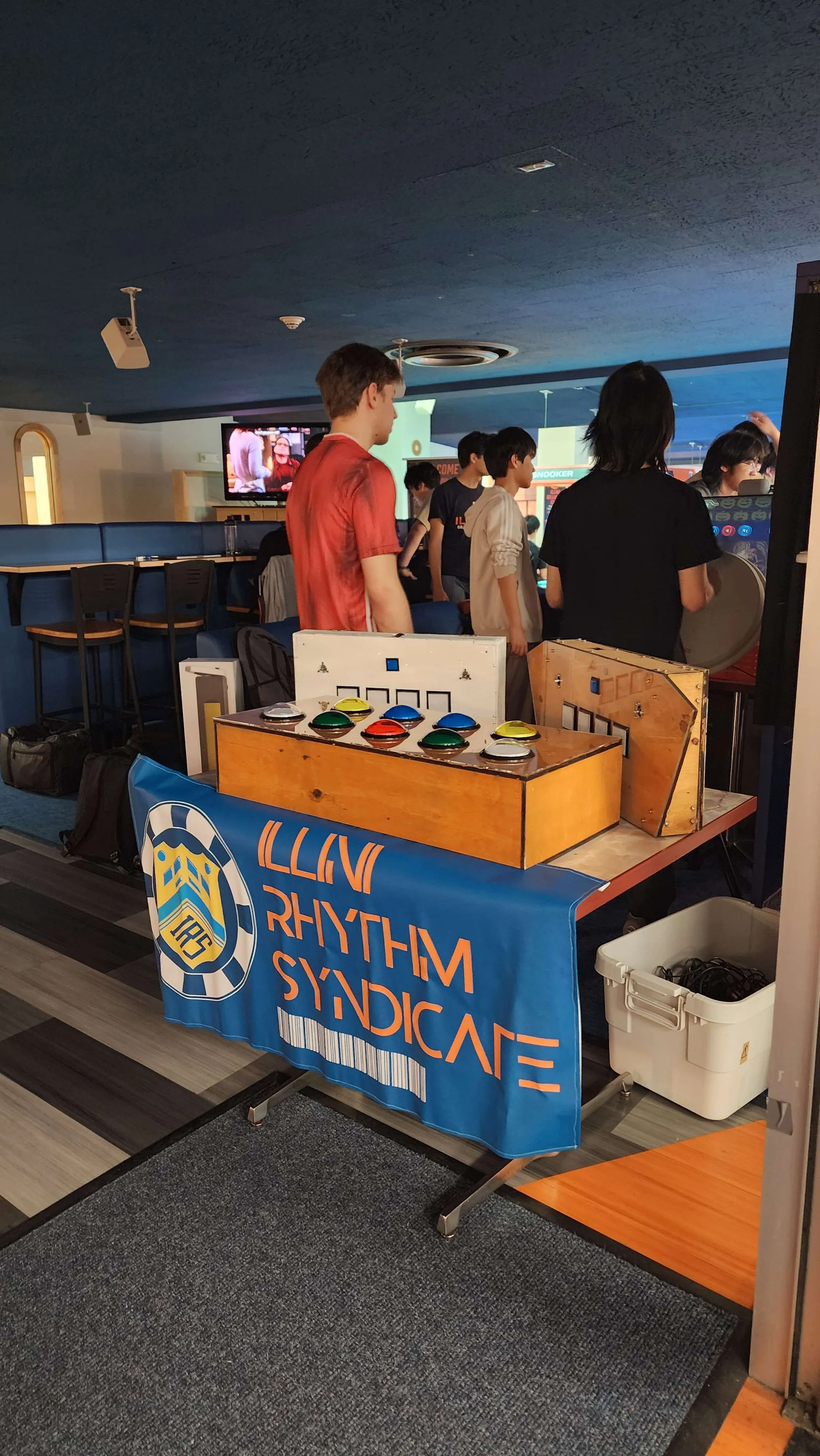 Two wooden club-built rhythm game controllers with colored arcade buttons set up on a table draped in an Illini Rhythm Syndicate banner