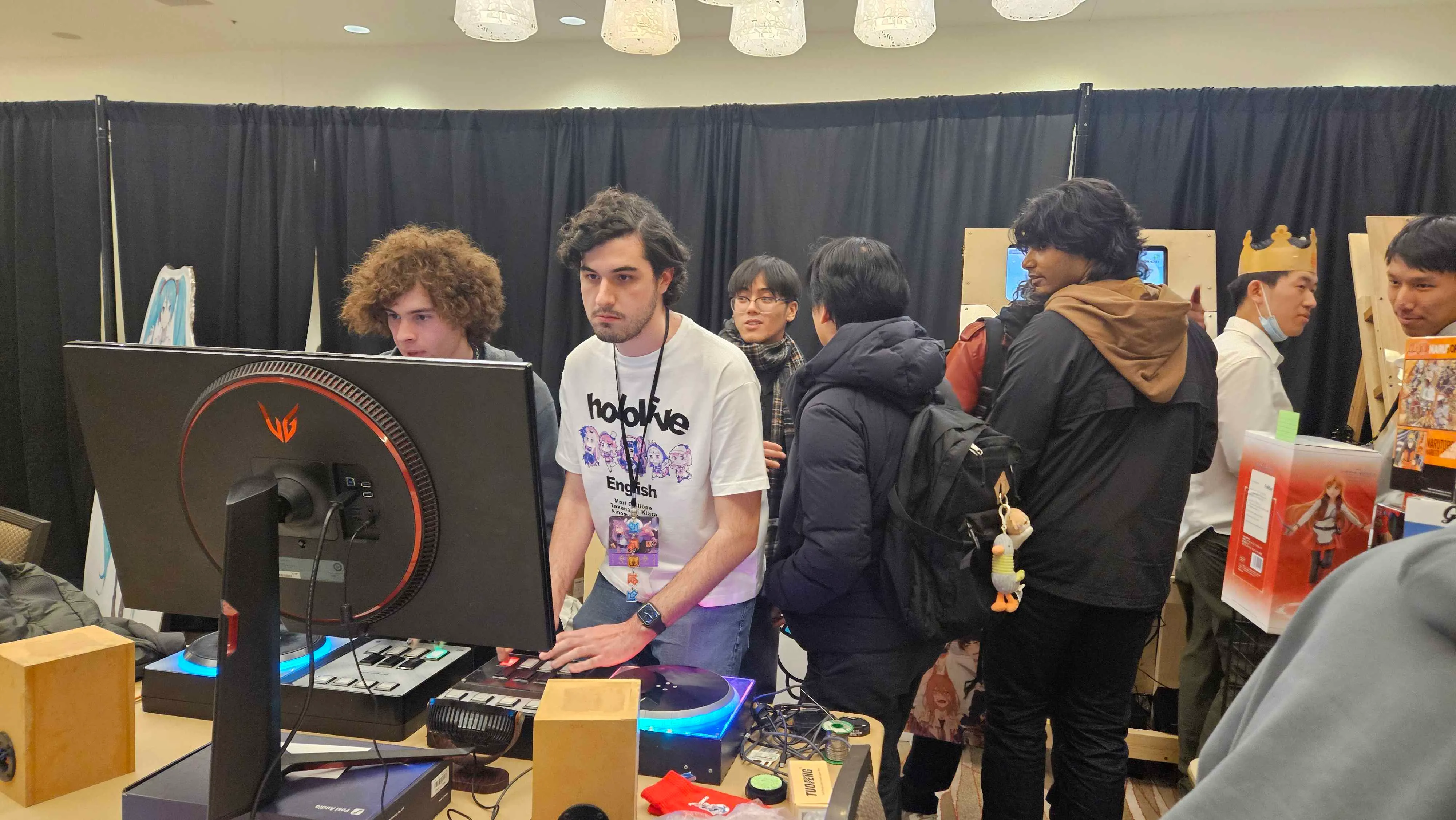 Members running a rhythm game setup with monitors and custom controllers at a UI-CON booth while attendees watch